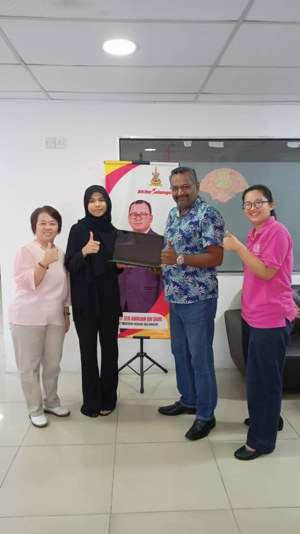 Four people standing indoors with a black laptop held between them